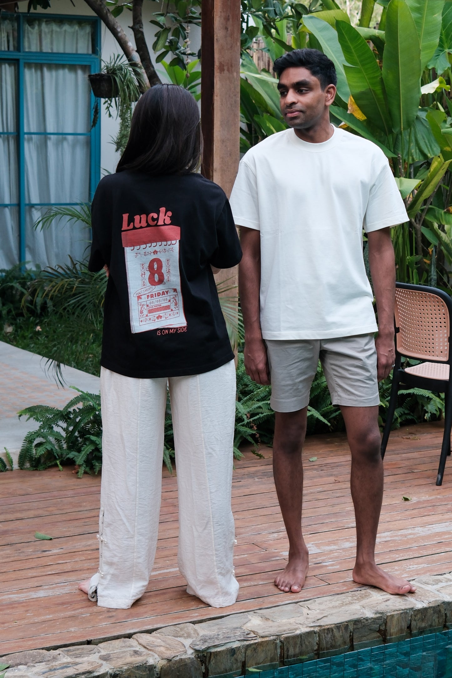 Two people wearing the 'Luck' T standing on a wooden deck with plants and a pool in the background