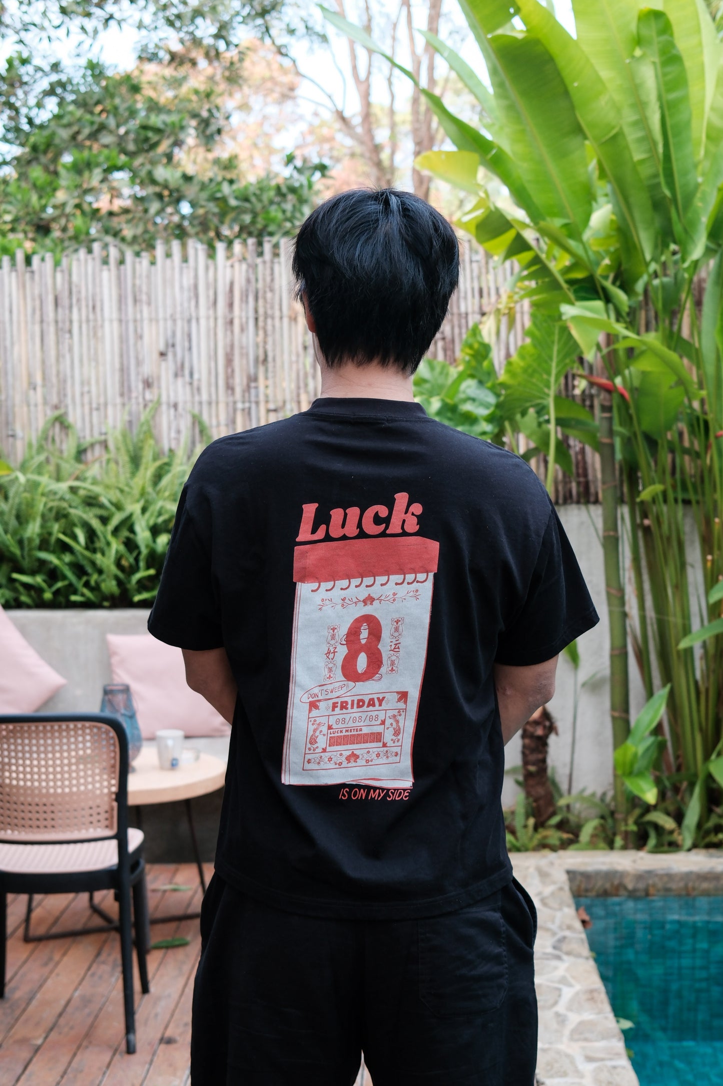 Man wearing a 'Luck' t-shirt standing outdoors near a pool and garden.